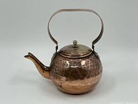 Front and side view of the vintage hammered copper kettle showing the rounded body, hinged lid with brass knob, and curved spout.