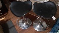 Photo showing pair of adjustable stools with black leather seats and chrome bases on a wooden floor.