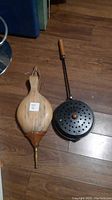 Two fireplace accessories placed on wooden floor: a wooden bellows with leather expanding section and brass nozzle, alongside a round cast iron tool with star-shaped perforations and wooden handle.
