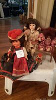 Photo showing four dolls: large German bisque doll in traditional red and black dress with hat, Victorian-style bisque doll, small clown doll seated on chair, miniature red dressed doll in clear case