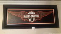 Framed Harley-Davidson emblem on a wall, showing black texture background with orange and white winged logo