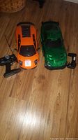 Two remote control cars side by side with their respective handheld controllers on a wooden floor.