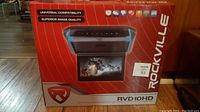 Front of the red retail box showing the Rockville brand and model RVD10HD. Image of the flip down screen showing a movie scene.