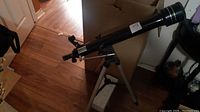 Black refractor telescope with tripod on wooden floor, angled to the side showing eyepiece and aiming scope