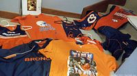 Several Denver Broncos clothing items laid out including T-shirts, jerseys, a little girls dress, and a book with sports theme.
