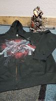 Black medium pull zip up hoodie with Gears of War 3 emblem and Marcus Fenix statue visible in background on carpet floor near wooden wall