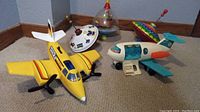 Photo showing all items including three spinning tops and two toy planes on a carpeted floor near baseboard corner.