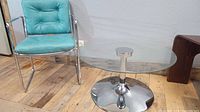 Teal green mid-century modern chair with chrome tubular frame and button tufted cushions shown with glass side table with chrome tulip base on wooden floor against white wall.