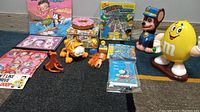 Wide view of the lot showing all items on a carpet including Betty Boop calendar, Garfield toy, Simpsons trivia game, and M&M dispenser