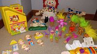 Wider view showing all items: dollhouse, Raggedy Ann dolls, Taco Bell chihuahuas, multiple troll dolls, and dog clown costume pieces on carpeted floor.