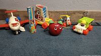 Five vintage Fisher Price pull toys arranged on blue carpet against wood background. Shows jet, kaleidoscope, apple, shoe, and dump truck.