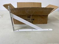 Photo showing a single white metal L-shaped shelf bracket with diagonal support bar against a white surface with open cardboard box behind it.