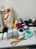 Top view showing full assortment including large cotton batting package, yarn skeins in gray and gradient colors, spools of thread, and orange scissors.