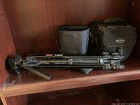 Optex T250 tripod folded with black Optex branded carrying bag and another black camera bag on wooden shelf