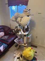 Upholstered beige cat tree with multiple levels, scratching posts, and plush surfaces. Various stuffed animals placed on and around it.