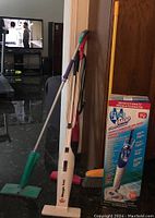 Photo of the five cleaning tools including the H2O steam mop in box, Bissell Magic Vac, 3-in-1 vacuum/broom, yellow broom and green flat mop arranged against a wall.