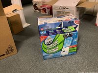 Photo of the lot showing the Scrubbing Bubbles Automatic Shower Cleaner sealed box and other boxed items in background on the floor.