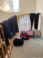 Photo showing various clothing items draped over and on floor near wood railing, including dark pants, scarves, white hooded pullover, and folded garments in pink, patterned, gray, and blue.