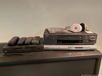 Front and angle view of Beats by Dre headphones, Toshiba VCR M-754C, Koss DVD player KS3122-2, RCA DirecTV box, and four remotes on top of older TV set.