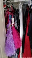Closet view showing an assortment of dresses in various colors including lavender, pink, white, black, and red with embellishments such as sequins, ruffles, and beading.