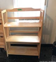 Front view of the wooden folding shelf unit showing four shelves and slatted side panels with a curved top back panel. Some stains and markings visible on the shelves.
