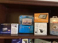 A shelf with boxed and packaged items including Sony Handycam DCR-SR47 camcorder, TomTom XL 330S GPS, Garmin DriveSmart 50 GPS, Garmin nüvi 1350 GPS, and a pedometer device.