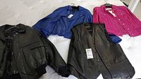 Four garments laid on a bed, showing black leather jacket, blue jacket, pink jacket, and a black vest