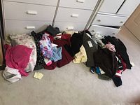 Photo of assorted folded ladies, girls, and mens clothing items including tops and pants on the floor in front of dressers.