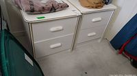 Pair of white laminate night stands side by side showing front and top surfaces, drawers closed, with some items on top