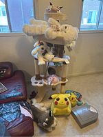 Shows plush cat tower with multiple levels holding several plush animals including a white teddy bear, small white dog, pink plush and two Pikachu toys on the floor next to it.