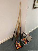 Full lot view showing six pool cues leaning against the wall, two boxes of pool balls, and the rack on the floor.