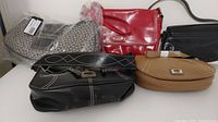 All five purses shown together on a white surface, includes black Guess purse, red purse, tan purse, black purse, and monogram purse in plastic with tags.