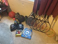 Overview of sports equipment including basketballs, football, racquets, frisbees, pads on floor against wall.