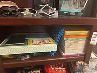 Shelf holding white and black sunglasses, black and white striped computer mouse, packs of office cards in a box, reams of colorful paper, and a blue plastic stand.