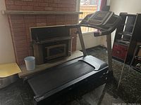 Side view of the treadmill showing running belt, gray support rails, and black frame next to a fireplace.