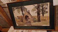 Framed watercolor artwork showing a rustic stone arch with a cross and trees around it, set against a wall and a building, photo taken to document condition and view of painting.