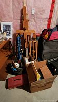 Photo showing two large wooden easels, one small wooden easel, blue metal easel, metal music stand, tripod, assorted art supplies stored in boxes and bags in basement setting.