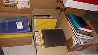 Three cardboard boxes filled with assorted books in various conditions and sizes on a basement floor with visible cardboard and appliance boxes in background