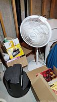 White Lasko oscillating floor fan on round base positioned near cardboard boxes and a black revolving step stool on floor.