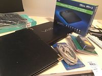 Lot items on table including black Vaio laptop, unopened router box, multimeter and cable in original packaging.