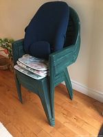 Four stacked dark green plastic patio chairs with armrests, with four floral patterned seat cushions stacked on top, and one large blue Obus Form back cushion and one small matching lumbar cushion leaning on the top chair.