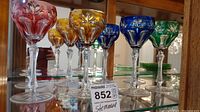 Photo showing the full set of 8 colored cut glass stemware with red, amber, blue, and green glasses arranged in a line on a glass shelf inside a wooden cabinet.