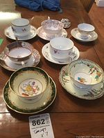 Seven assorted sets of teacups and saucers arranged on table showing variety of floral and patterned designs with some gold accents.