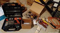 Photo of assorted tools including a Black & Decker cordless drill in a black plastic carrying case, boxes with various hand tools, pliers, saws, and an orange extension cord