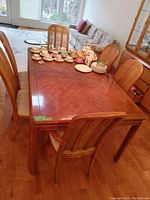 Angle view of the vintage dining table with six chairs and multiple tea set pieces on top. The wooden table shows a rich brown finish with wood grain patterns, surrounded by six cane back chairs, one with arms.