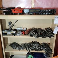 Photo showing several Lionel train cars including black locomotive, red caboose, tender car, and metal tracks stacked on shelves.