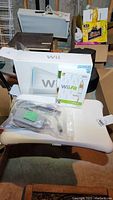 Nintendo Wii console with original box, controllers, cables, and Wii Fit game disc on white chair.