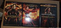 Three DVD cases of The Hunger Games film series lying on a cardboard surface. Cases include Mockingjay Part 1, Mockingjay Part 2, and a 2-movie set with Catching Fire and The Hunger Games.