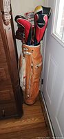 Photo of the full golf bag filled with clubs, standing upright against a door and furniture, showing tan leather bag and some club headcovers.