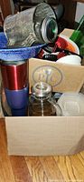Photo showing assorted kitchen items in a cardboard box including galvanized metal flour sifter, blue stoneware dish, insulated mugs, plastic containers, glass carafe with metal lid, and various utensils.